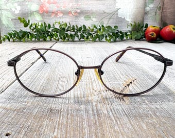 May include: Oval spectacles with a dark brown metal frame. The lenses are clear with a slight yellow tint. The temples have a textured design. The glasses are set against a rustic wooden background.