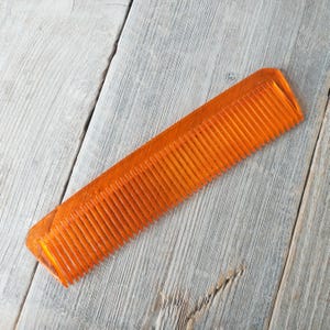 May include: An orange, rectangular comb with fine teeth. The comb is made of a translucent material and is set against a weathered, gray wooden surface. The comb's teeth are evenly spaced.