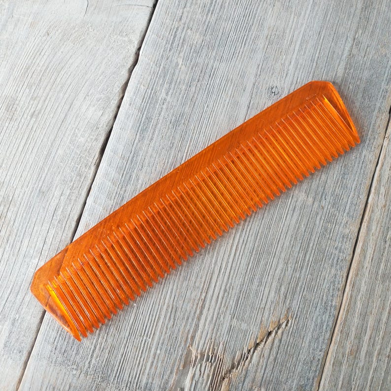May include: An orange, rectangular comb with fine teeth. The comb is made of a translucent material and is set against a weathered, gray wooden surface. The comb's teeth are evenly spaced.