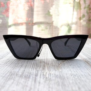 Puede incluir: Gafas de sol negras con una forma de ojo de gato angular y afilada. Las monturas son brillantes y las lentes oscuras, proporcionando un aspecto elegante y moderno. Las gafas de sol se muestran sobre una superficie de madera clara.