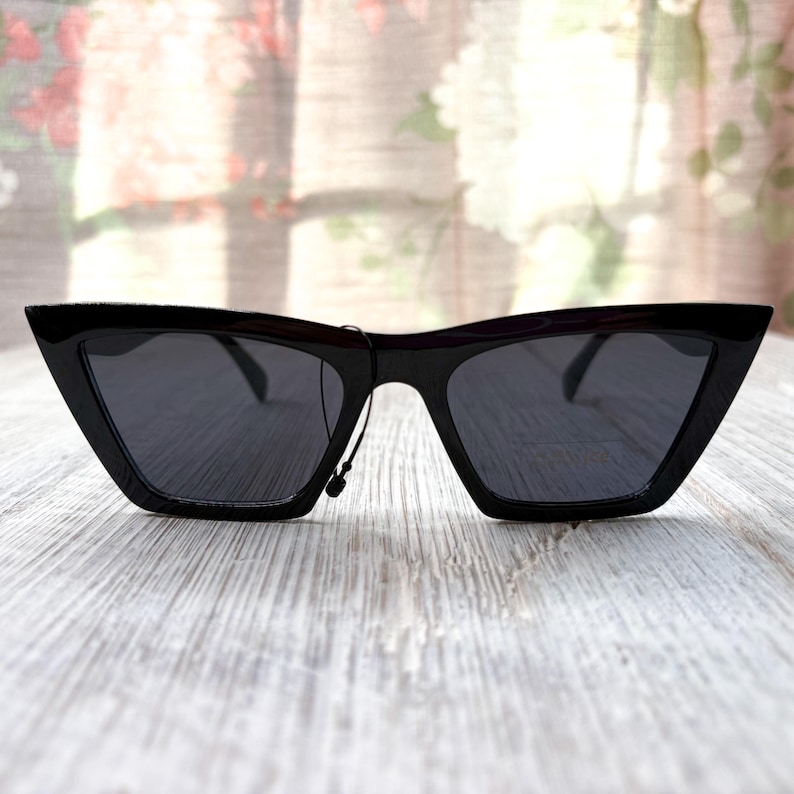 Puede incluir: Gafas de sol negras con una forma de ojo de gato angular y afilada. Las monturas son brillantes y las lentes oscuras, proporcionando un aspecto elegante y moderno. Las gafas de sol se muestran sobre una superficie de madera clara.