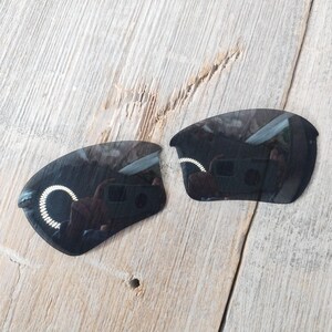 Op de afbeelding: Twee zwarte vervangende glazen voor zonnebrillen. De glazen zijn gebogen en hebben een donkere tint.