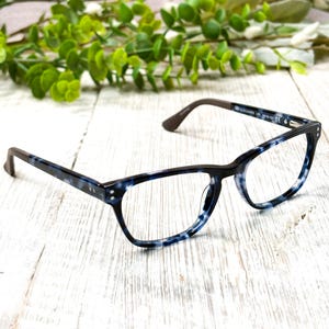 May include: Eyeglasses with a blue and white speckled pattern on the frames and dark brown arms. The glasses are set against a white wooden surface with green foliage in the background.