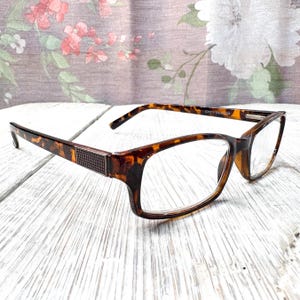 May include: Tortoiseshell-patterned eyeglasses with a rectangular frame. The glasses have clear lenses and a decorative metal accent on the temples. The arms of the glasses are also tortoiseshell.