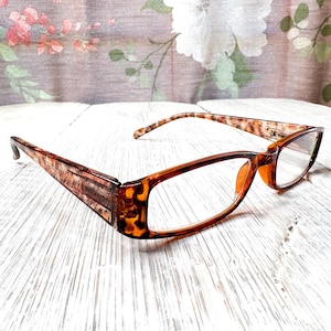 Könnte beinhalten: Eine Brille mit braunen Schildpatt-Fassungen und Bügeln. Die Gläser sind klar. Sie liegen auf einer weißen Holzoberfläche. Der Hintergrund zeigt einen Stoff mit floralem Muster.