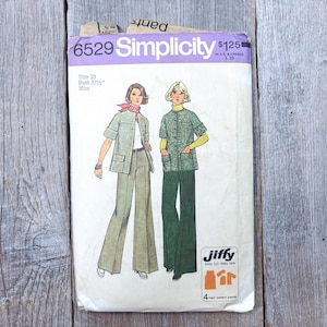 Könnte beinhalten: Vintage Simplicity Schnittmuster 6529, mit einem zweiteiligen Hosenanzug-Design. Der Musterumschlag zeigt Illustrationen des Anzugs, mit einer Jacke und Schlaghose. Der Umschlag befindet sich auf einer verwitterten Holzoberfläche.