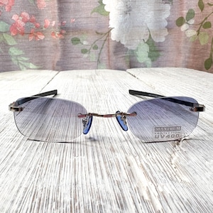 May include: Rimless sunglasses with a gradient blue tint. The glasses have a silver bridge and hinges, with black arms. The lenses have the text "MAXIMUM PROTECTION UV400". The sunglasses are on a white wooden surface.