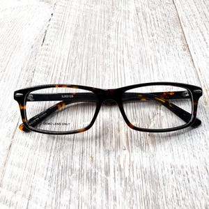 May include: Rectangular eyeglasses with a tortoiseshell pattern frame. The glasses have a black bridge and temples. The text "DEMO LENS ONLY" is visible on the frame. The glasses are on a white wooden surface.