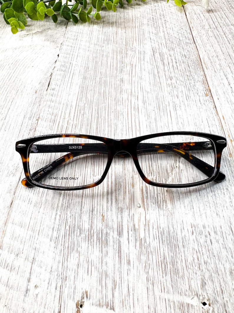 May include: Rectangular eyeglasses with a tortoiseshell pattern frame. The glasses have a black bridge and temples. The text "DEMO LENS ONLY" is visible on the frame. The glasses are on a white wooden surface.