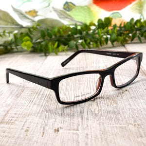May include: Black rectangular eyeglasses with clear demo lenses. The frames have a glossy finish and a contrasting red accent on the inner arms. The text "DEMO LENS ONLY" is visible on the lenses.