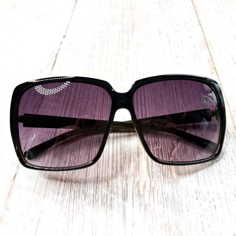 以下が含まれることがあります： 黒いスクエアフレームのサングラス、紫色のグラデーションレンズ付き。 サングラスはフレーム上部に装飾的な要素があります。 メガネは明るい木目調の背景に置かれています。