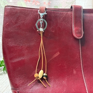 May include: A red leather tote bag with a silver keychain and a decorative tassel. The tassel features wooden beads and leather strands. The bag has a handle and white stitching.