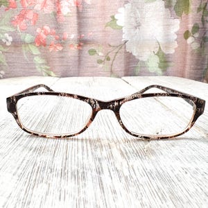 May include: Eyeglasses with a brown and white patterned frame. The rectangular lenses are clear. The glasses are set against a background of a floral pattern on a light-colored surface.