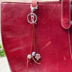 May include: A red leather tote bag with a silver-tone keychain and a decorative tassel. The tassel features brown leather strands, two decorative beads, and a flower-shaped charm. The bag has a handle and a strap.