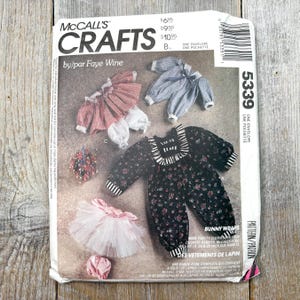 May include: A vintage McCall's Crafts sewing pattern envelope featuring illustrations of bunny clothing. The pattern includes designs for various outfits, including a floral jumpsuit, a tutu, and striped tops. The text on the envelope reads "Bunny Wraps."