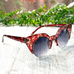 May include: Red and white patterned sunglasses with a cat-eye shape and gradient lenses. The sunglasses have a unique design and are set against a background of greenery and a floral pattern.
