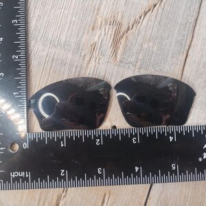 May include: Two black replacement lenses for sunglasses, measured against a ruler. The lenses are approximately 2 inches wide.