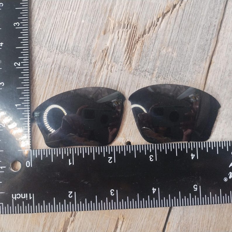 May include: Two black replacement lenses for sunglasses, measured against a ruler. The lenses are approximately 2 inches wide.