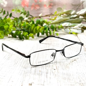 May include: A pair of rectangular eyeglasses with a dark frame and clear demo lenses. The text "DEMO LENS ONLY" is printed on one lens. The glasses are set against a white wooden surface with green foliage and pink flowers in the background.