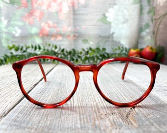 May include: Round-framed spectacles in a reddish-brown colour. The glasses feature a classic design with a keyhole bridge and slender arms. The lenses are clear, and the frames have a glossy finish. The spectacles are placed on a light-coloured wooden surface.