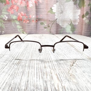May include: Eyeglasses with a dark brown metal frame and clear lenses. The frame has a half-rim design. The glasses are on a white wooden surface. The background has a floral pattern.