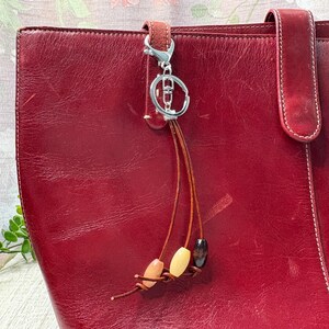 May include: A red leather tote bag with a silver keyring and a decorative tassel. The tassel has three wooden beads in shades of brown, cream, and black. The bag has a handle and a strap.