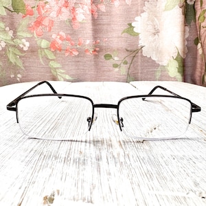 Puede incluir: Un par de gafas con montura de metal marrón oscuro. Las lentes son transparentes y rectangulares, con un diseño de media montura. Las gafas están sobre una superficie de madera blanca, con una cortina floral.
