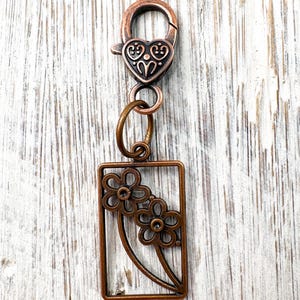 May include: A copper-colored keychain with a heart-shaped clasp and a rectangular charm featuring two stylized flowers. The keychain is made of metal and has a vintage or antique appearance.