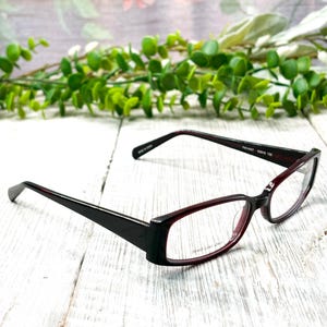 Puede incluir: Gafas rectangulares con montura negra y detalles burdeos. Las lentes son transparentes y la montura tiene un diseño elegante y moderno. Las gafas están sobre una superficie de madera blanca con follaje verde en el fondo.