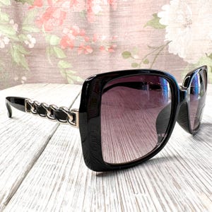 May include: Black square sunglasses with gradient lenses. The frames are thick and glossy, with a chain detail on the temples. The lenses transition from a dark purple at the top to a lighter shade at the bottom.