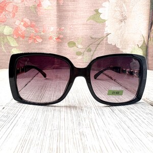 Puede incluir: Gafas de sol negras de gran tama&ntilde;o con lentes degradadas. Las gafas tienen una montura rectangular y una peque&ntilde;a pegatina que dice "UV 400". Las gafas est&aacute;n sobre una superficie de madera blanca.