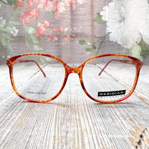 May include: Vintage eyeglasses with a tortoiseshell pattern. The frames are a warm blend of orange, red, and yellow. The lenses are clear, and the text "DEMO LENS ONLY" and "MERIDIAN COLLECTION" are visible. The glasses have a classic, oversized shape.