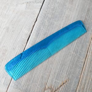 May include: A translucent blue comb with fine teeth. The comb is made of a solid, durable material and has a rectangular shape with rounded edges. The comb is angled across a light gray wooden surface.