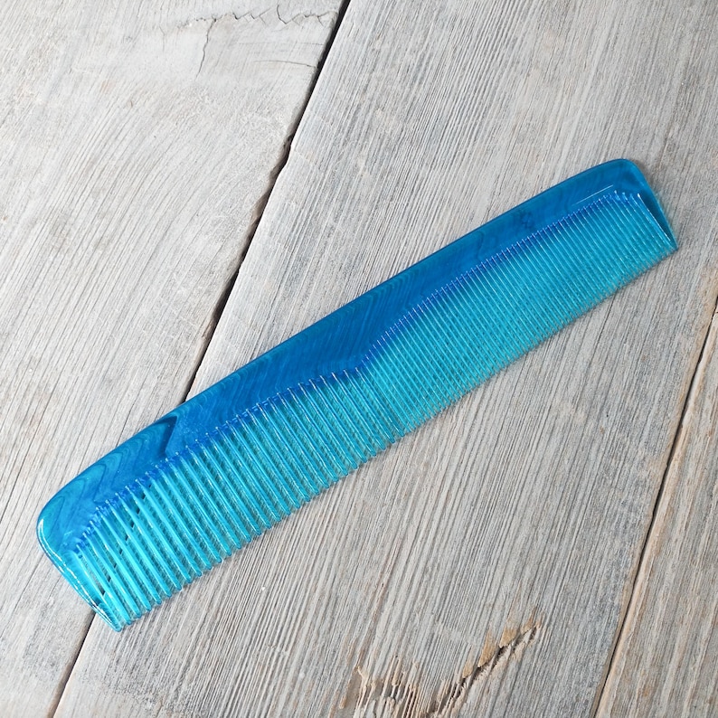 May include: A translucent blue comb with fine teeth. The comb is made of a solid, durable material and has a rectangular shape with rounded edges. The comb is angled across a light gray wooden surface.