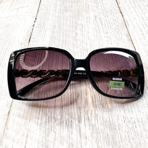 Puede incluir: Gafas de sol cuadradas negras con detalle de cadena en las patillas y lentes degradadas. Las gafas de sol tienen una pegatina verde que dice "MAXIMUM UV 400 UV PROTECTION".