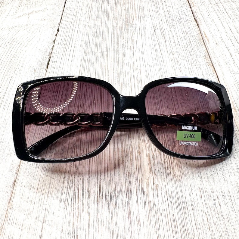 Puede incluir: Gafas de sol cuadradas negras con detalle de cadena en las patillas y lentes degradadas. Las gafas de sol tienen una pegatina verde que dice "MAXIMUM UV 400 UV PROTECTION".