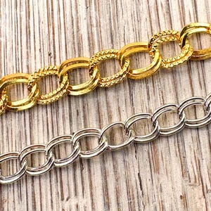 May include: Close-up of two metal chain links. One chain is gold-colored with a textured design, and the other is silver-colored. The chains are on a light brown wooden surface.