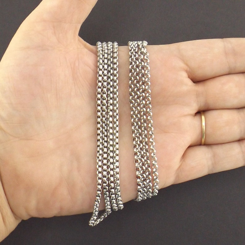 May include: Two silver chains, one thicker than the other, are laid out on a hand. The chains are made of small, linked circles.