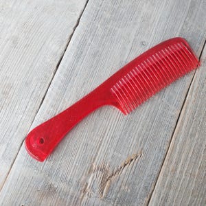 May include: A red plastic comb with a handle and fine teeth. The comb is designed for hair grooming and styling. The handle has a hole for hanging. The comb is shown on a weathered wooden surface.