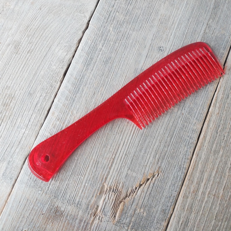 May include: A red plastic comb with a handle and fine teeth. The comb is designed for hair grooming and styling. The handle has a hole for hanging. The comb is shown on a weathered wooden surface.