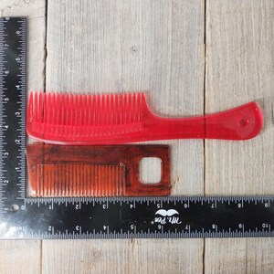 May include: Two combs are displayed on a wooden surface. A transparent red comb with a handle is positioned above a smaller, rectangular brown comb. A ruler is placed next to the combs for scale. The combs are for hair care.