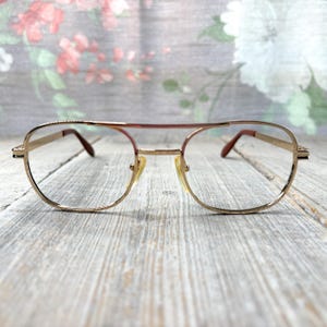 May include: Gold-coloured eyeglasses with a double bridge and clear lenses. The frames have a classic aviator style with a thin, rectangular shape. The temples are a dark brown colour, and the background is a floral pattern.