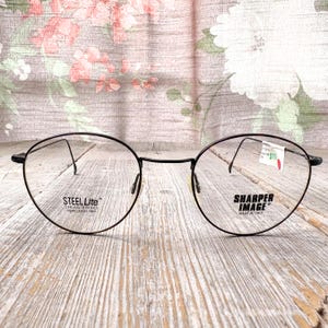 May include: Black round eyeglasses with thin frames. The left lens has the text "STEEL Lite" and the right lens has the text "SHARPER IMAGE MADE IN ITALY". The glasses have spring hinges.
