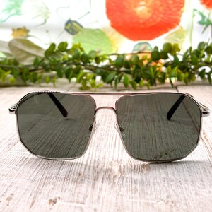 Pode incluir: &Oacute;culos de sol com arma&ccedil;&atilde;o prateada e lentes escuras. Os &oacute;culos t&ecirc;m formato geom&eacute;trico e dupla ponte. As lentes s&atilde;o de cor cinza escuro. Os &oacute;culos de sol est&atilde;o sobre uma superf&iacute;cie de madeira clara.