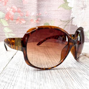 Peut inclure: Lunettes de soleil marron à motif écaille de tortue avec monture surdimensionnée. Les lunettes ont des verres dégradés et des détails dorés sur les branches. Style rétro classique.