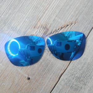 May include: Two blue mirrored replacement lenses for sunglasses. The lenses are curved and have a reflective blue coating.