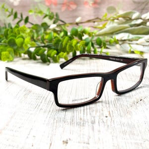 May include: Black and brown rectangular eyeglasses with clear lenses. The frames have a glossy finish and a contrasting brown interior. The text "DEMO LENS ONLY" is visible on the lower frame. The glasses are set against a white wooden surface.