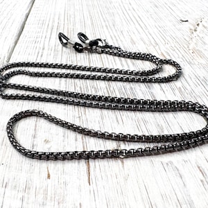 以下が含まれることがあります： ガンメタルグレーのメガネチェーン、ボックスチェーンのデザイン。 チェーンは、メガネに固定するための黒いラバーループに接続されています。 チェーンは白い木製の表面に置かれています。