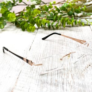May include: Two pairs of eyeglasses with clear lenses and rose gold frames are displayed on a white wooden surface. One pair has black temples with a decorative accent, while the other has a minimalist design. The text "Stainless Steel" is visible.