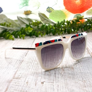 May include: White sunglasses with a geometric pattern on the top of the frames. The sunglasses have dark lenses and a white frame. The arms are white with a gold accent. The sunglasses are on a white wooden surface.